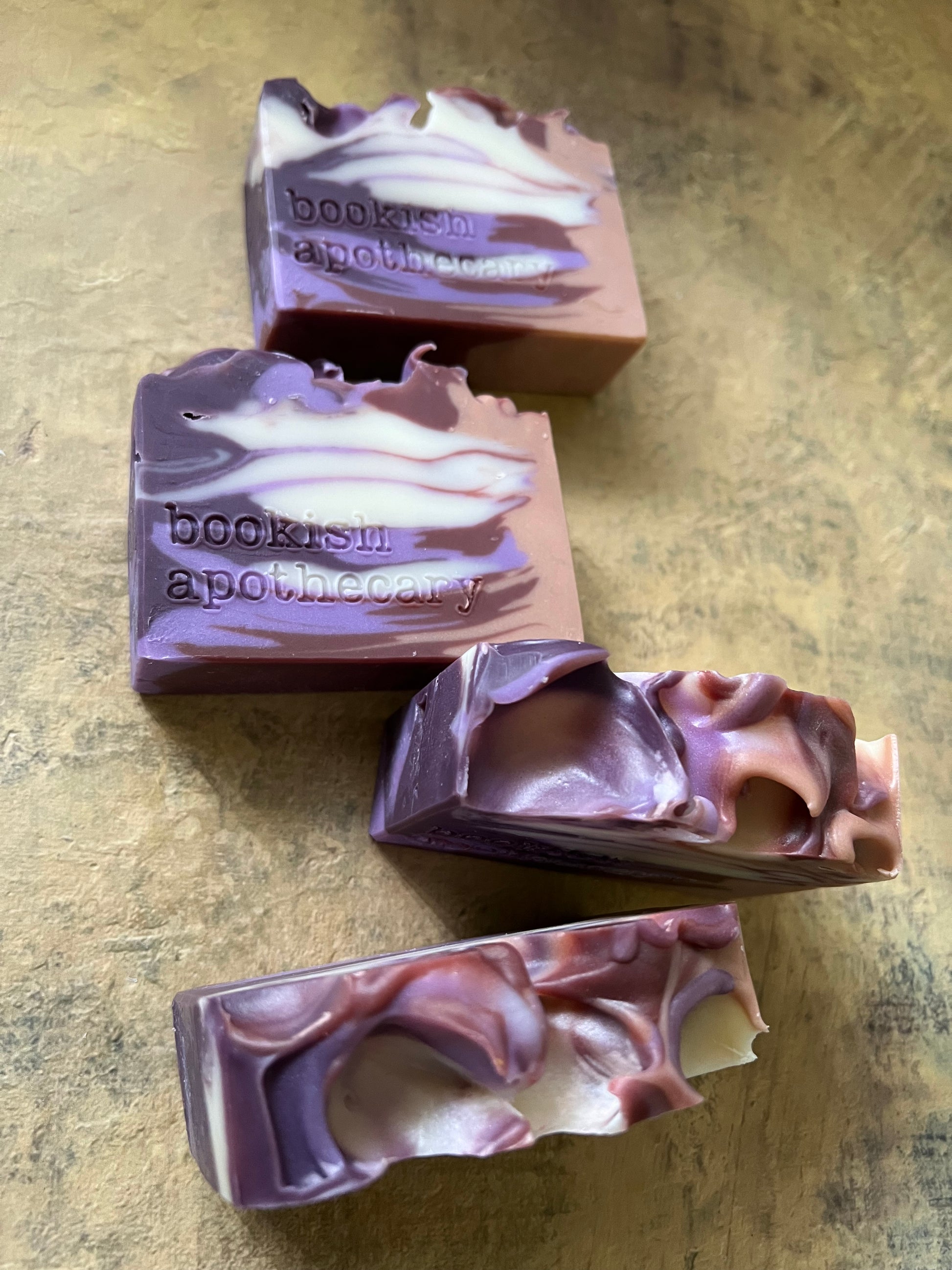 Four handmade soap bars with purple, brown, and white marbled swirls, featuring the stamped brand name 'bookish apothecary', arranged on a textured brown surface.