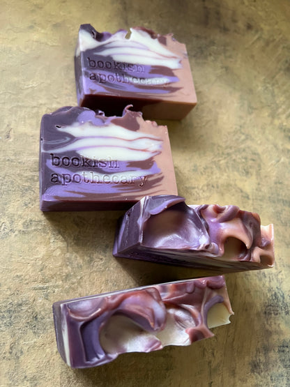 Four handmade soap bars with purple, brown, and white marbled swirls, featuring the stamped brand name 'bookish apothecary', arranged on a textured brown surface.