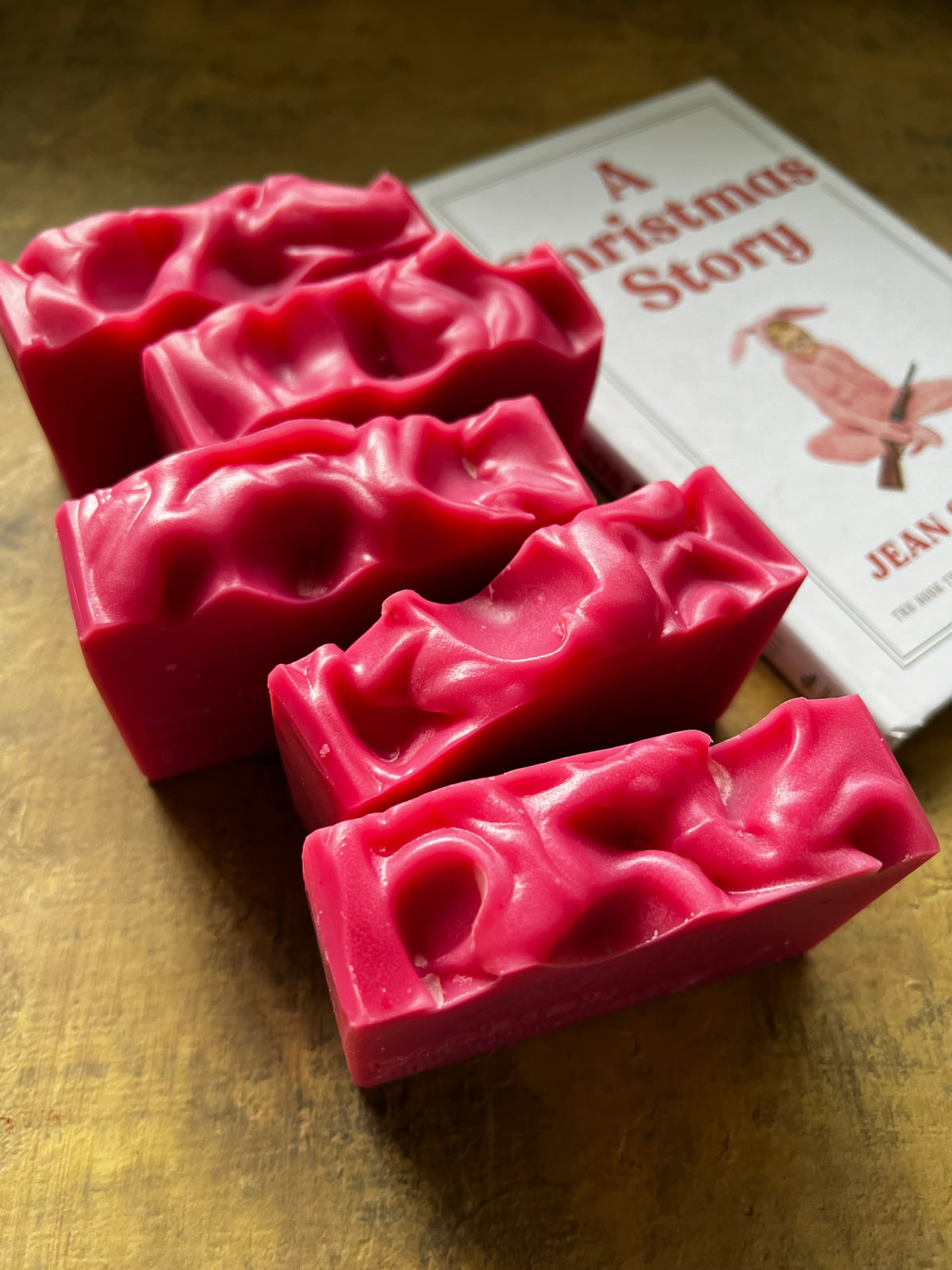 Five bright pink textured rectangular bars arranged on a golden surface next to a book titled "A Christmas Story."