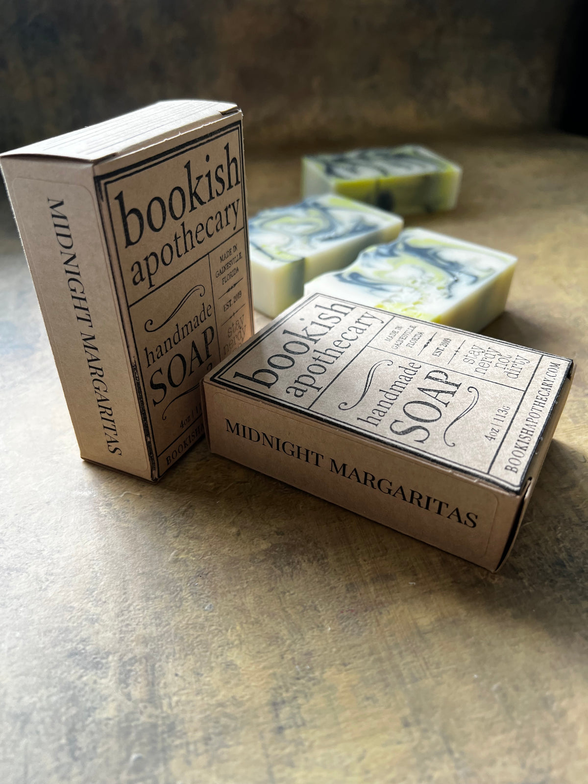 Two boxed bars of handmade soap from Bookish Apothecary labeled Midnight Margaritas, with two marbled soap bars visible behind them on a textured stone-like surface.