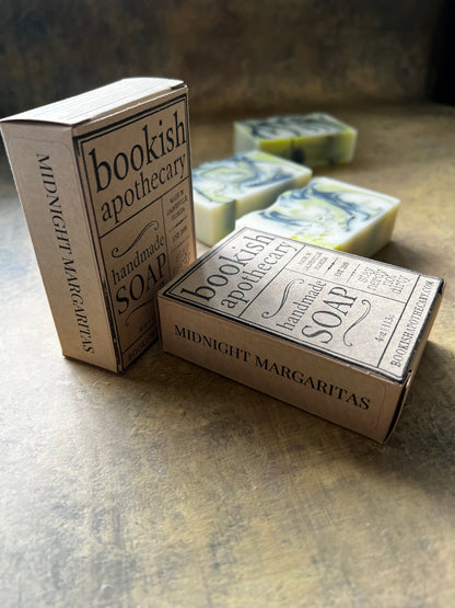 Two boxed bars of handmade soap from Bookish Apothecary labeled Midnight Margaritas, with two marbled soap bars visible behind them on a textured stone-like surface.