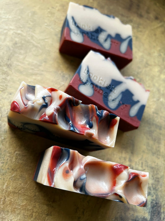 Four artisanal soap bars with a marbled pattern of cream, red, blue, and black, arranged diagonally on a textured gold surface.