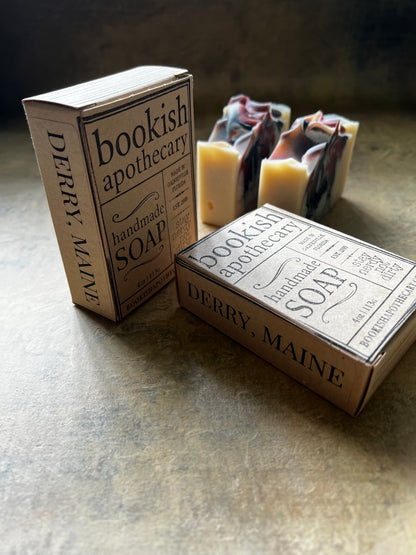 Two handmade soap bars with cream-colored soaps and dark swirls, displayed next to their vintage-style paperboard boxes printed with 'bookish apothecary' and 'DERRY, MAINE'.