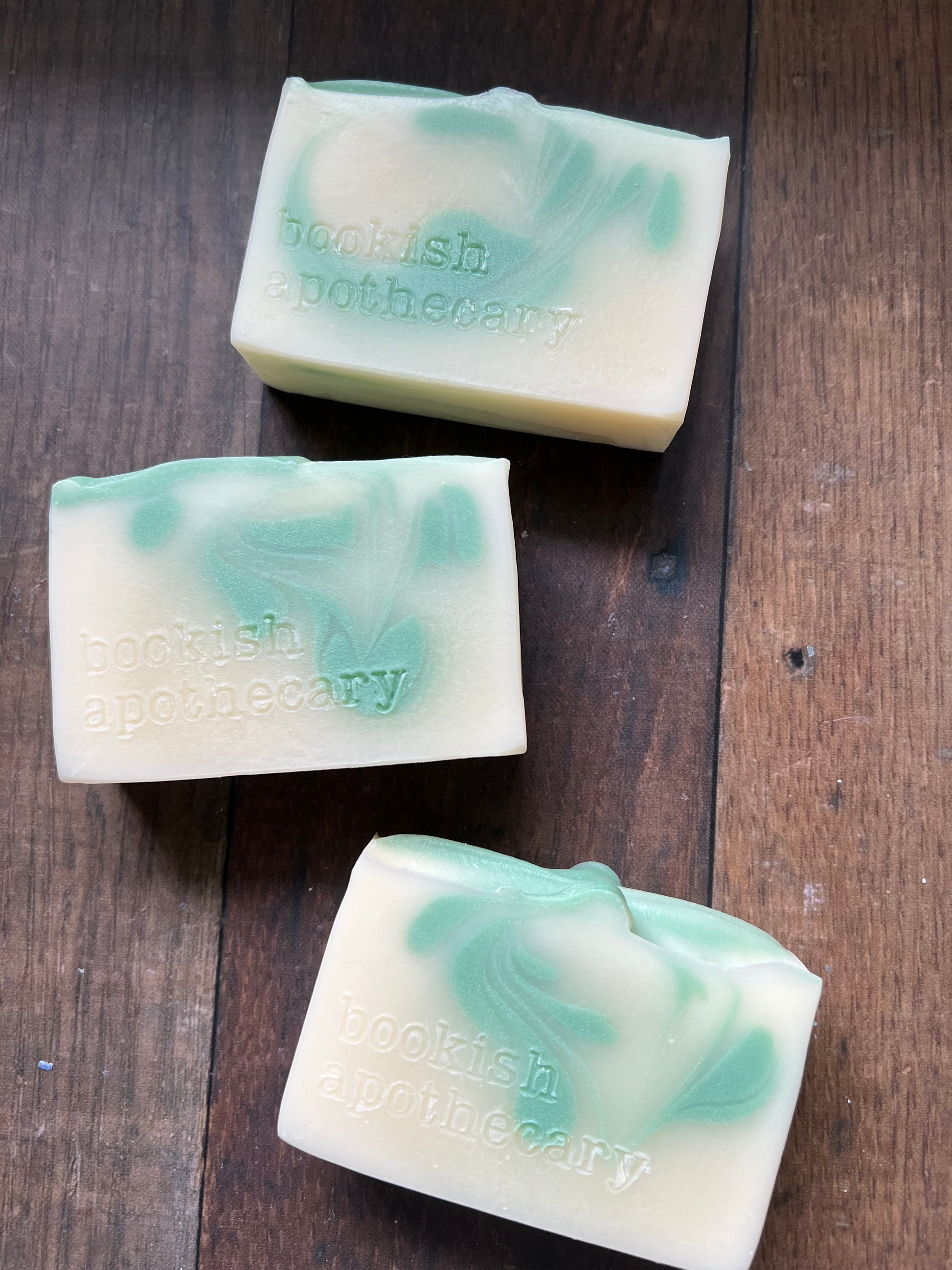 Three bars of white soap with green swirls stamped with "bookish apothecary" on a wooden surface