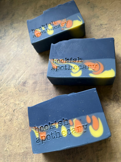 Three handmade soap bars with a dark blue-gray base and yellow and orange swirls, featuring the stamped text 'bookish apothecary' on a wooden surface.