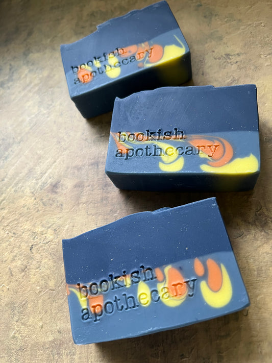 Three handmade soap bars with a dark blue-gray base and yellow and orange swirls, featuring the stamped text 'bookish apothecary' on a wooden surface.