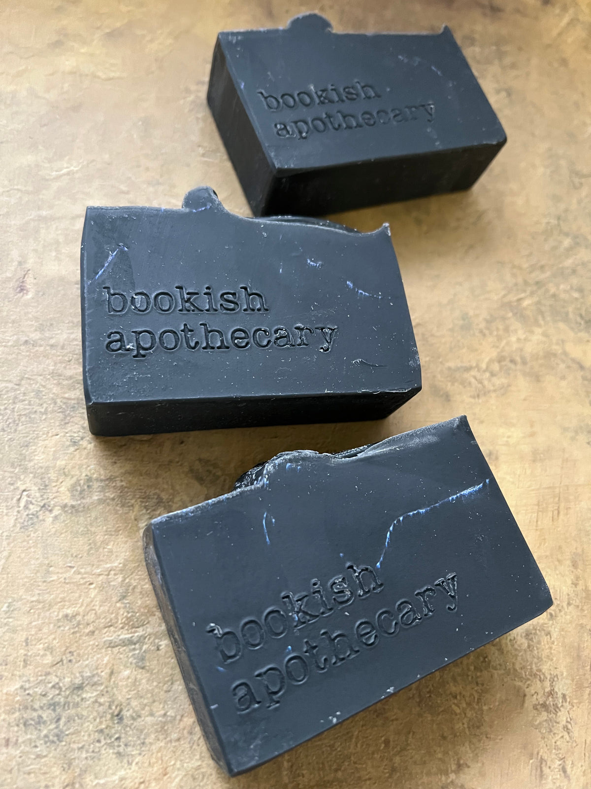 Three dark charcoal soap bars embossed with 'bookish apothecary' on a textured beige surface.