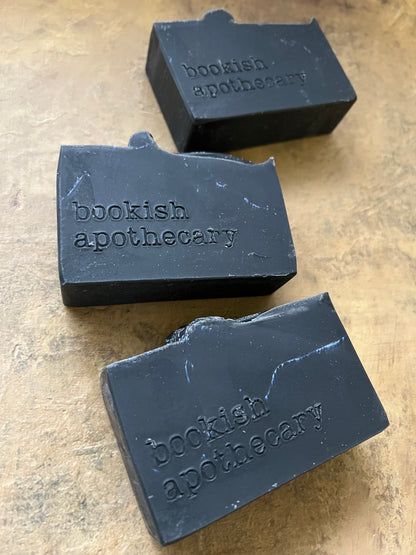 Three dark charcoal soap bars embossed with 'bookish apothecary' on a textured beige surface.