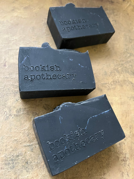 Three dark charcoal soap bars embossed with 'bookish apothecary' on a textured beige surface.