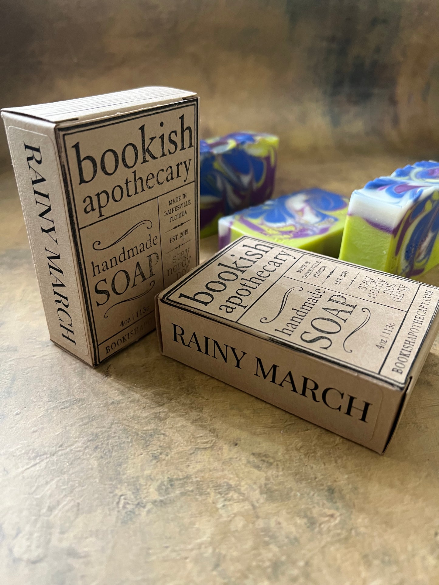 Two rectangular soap boxes labeled 'bookish apothecary handmade soap RAINY MARCH' on a textured beige surface, with marbled blue, green, and white soap bars partially visible behind them.