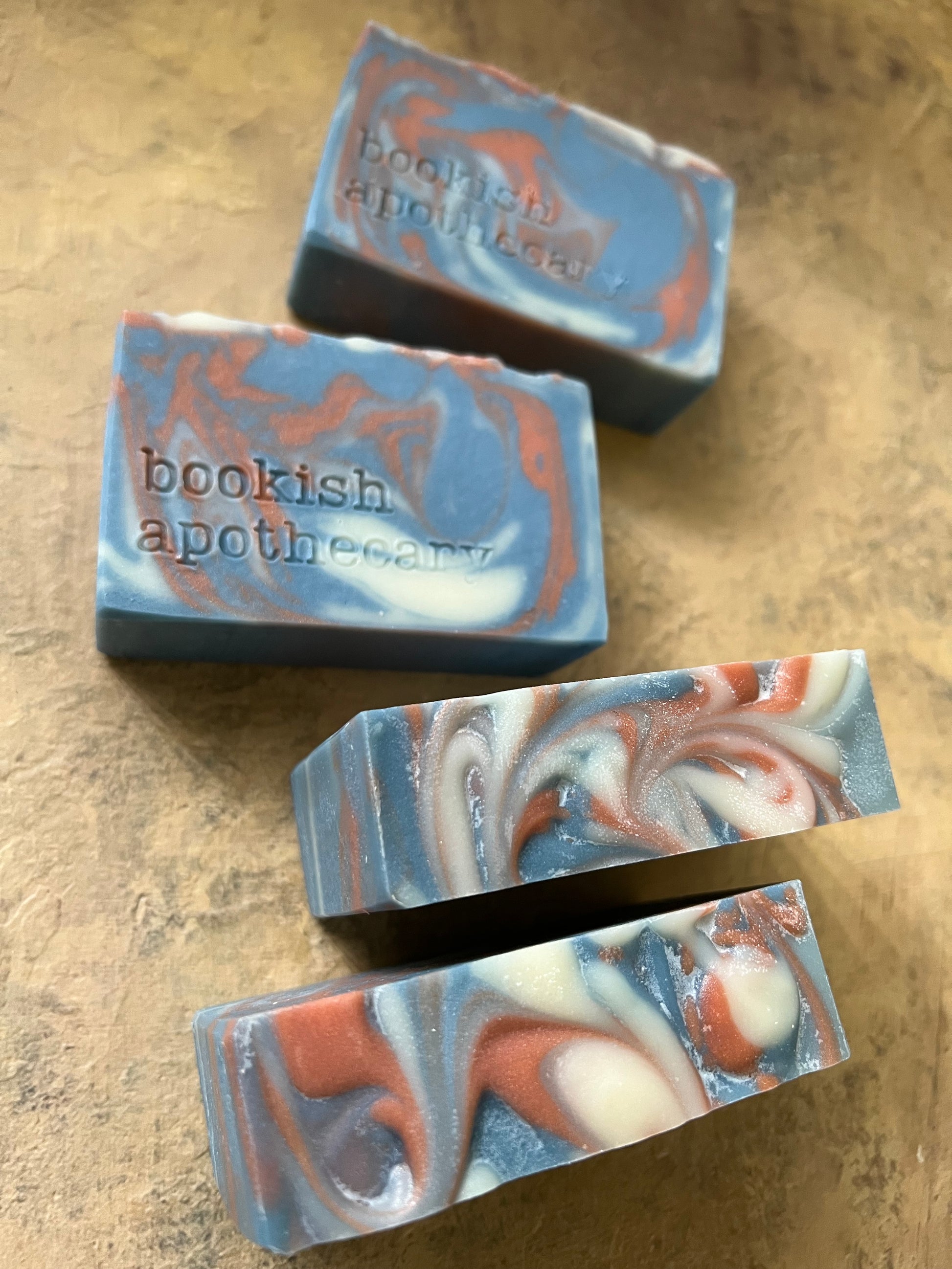 Four handmade soap bars with a swirled blue, white, and terracotta orange pattern,   'bookish apothecary' stamped on the soap, arranged on a textured beige surface.