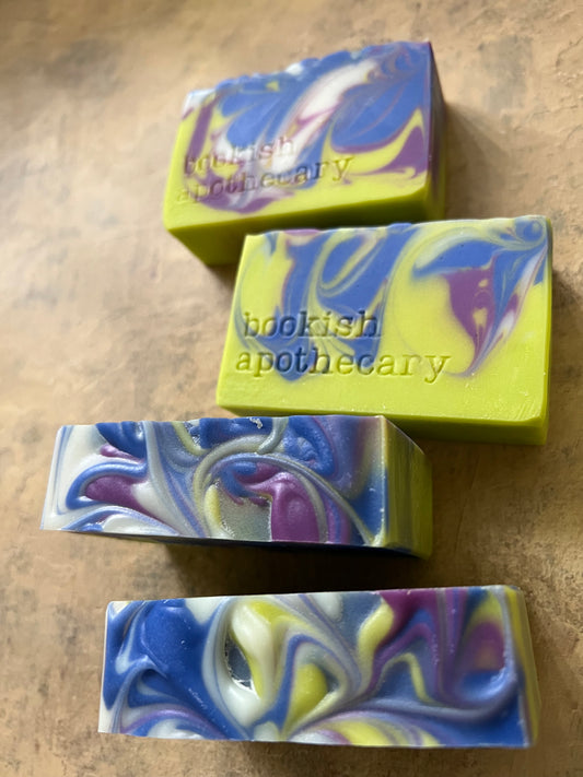 Four handmade marbled soap bars with blue, purple, yellow, and white swirls, arranged on a textured beige surface. The brand name 'bookish apothecary' is stamped on the bars.