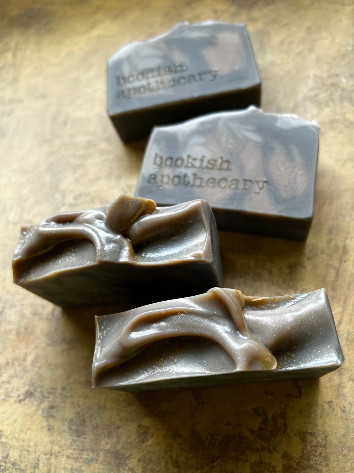Four rectangular bars of handmade soap with swirled dark brown and light tan patterns, embossed with the brand name 'bookish apothecary', arranged on a textured gold-toned surface.