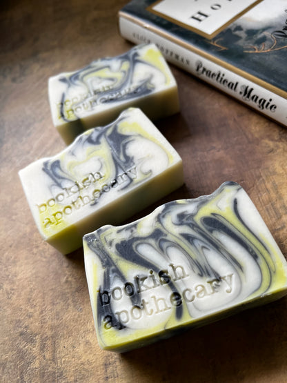 Three marbled soap bars with a white, gray, and yellow swirl pattern, embossed with the text 'bookish apothecary', arranged on a rustic wooden surface next to a book.