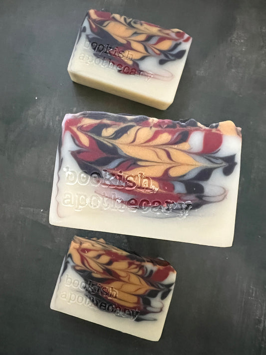 Three handmade soap bars with a marbled pattern of cream, red, yellow, and black, displayed on a dark gray surface.