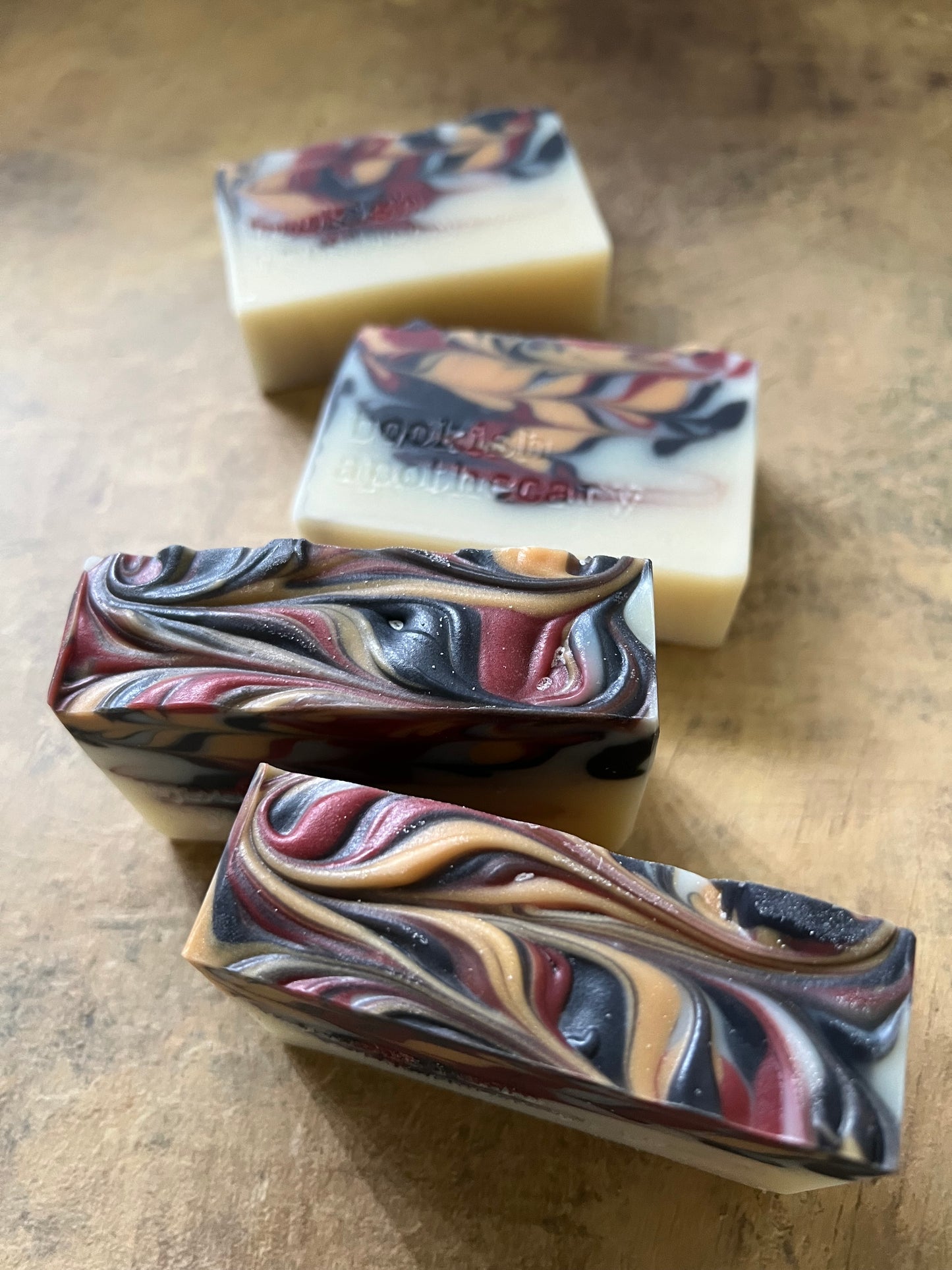 Four handmade marbled soap bars arranged diagonally on a textured brown surface. The soaps feature swirls of cream, burgundy, black, and tan.