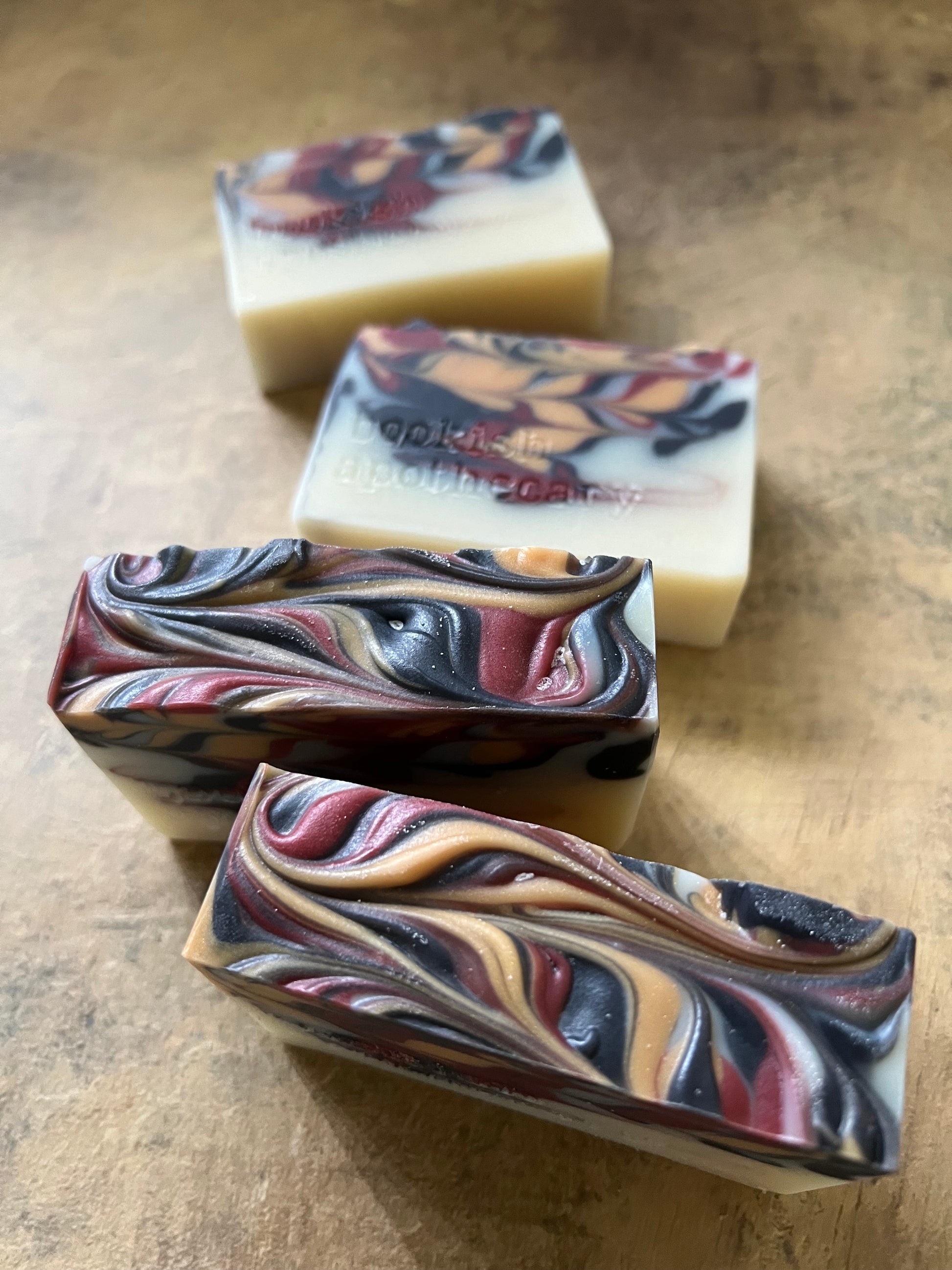 Four handmade marbled soap bars arranged diagonally on a textured brown surface. The soaps feature swirls of cream, burgundy, black, and tan.