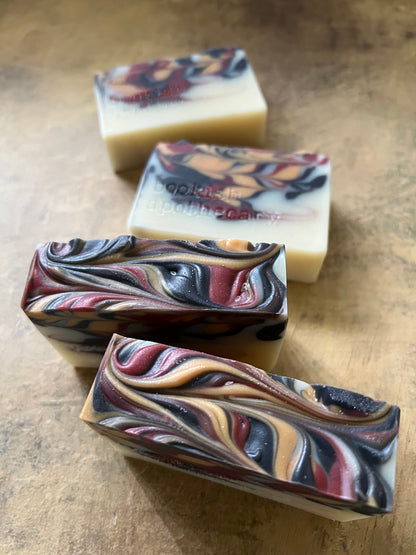 Four handmade marbled soap bars arranged diagonally on a textured brown surface. The soaps feature swirls of cream, burgundy, black, and tan.