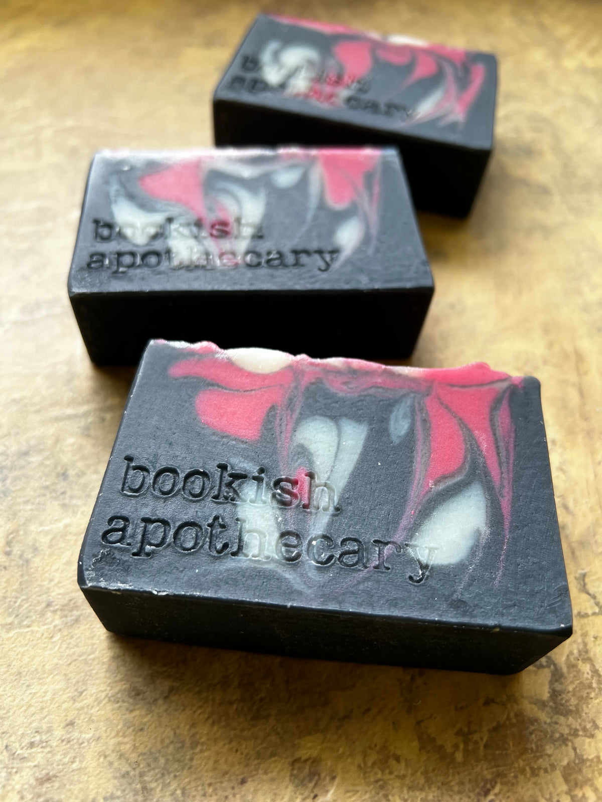 Three handmade soap bars with a black base and red, white, and gray marbled swirls, embossed with the text 'bookish apothecary', arranged on a textured brown surface.