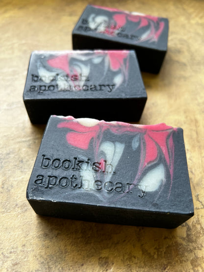 Three handmade soap bars with a black base and red, white, and gray marbled swirls, embossed with the text 'bookish apothecary', arranged on a textured brown surface.