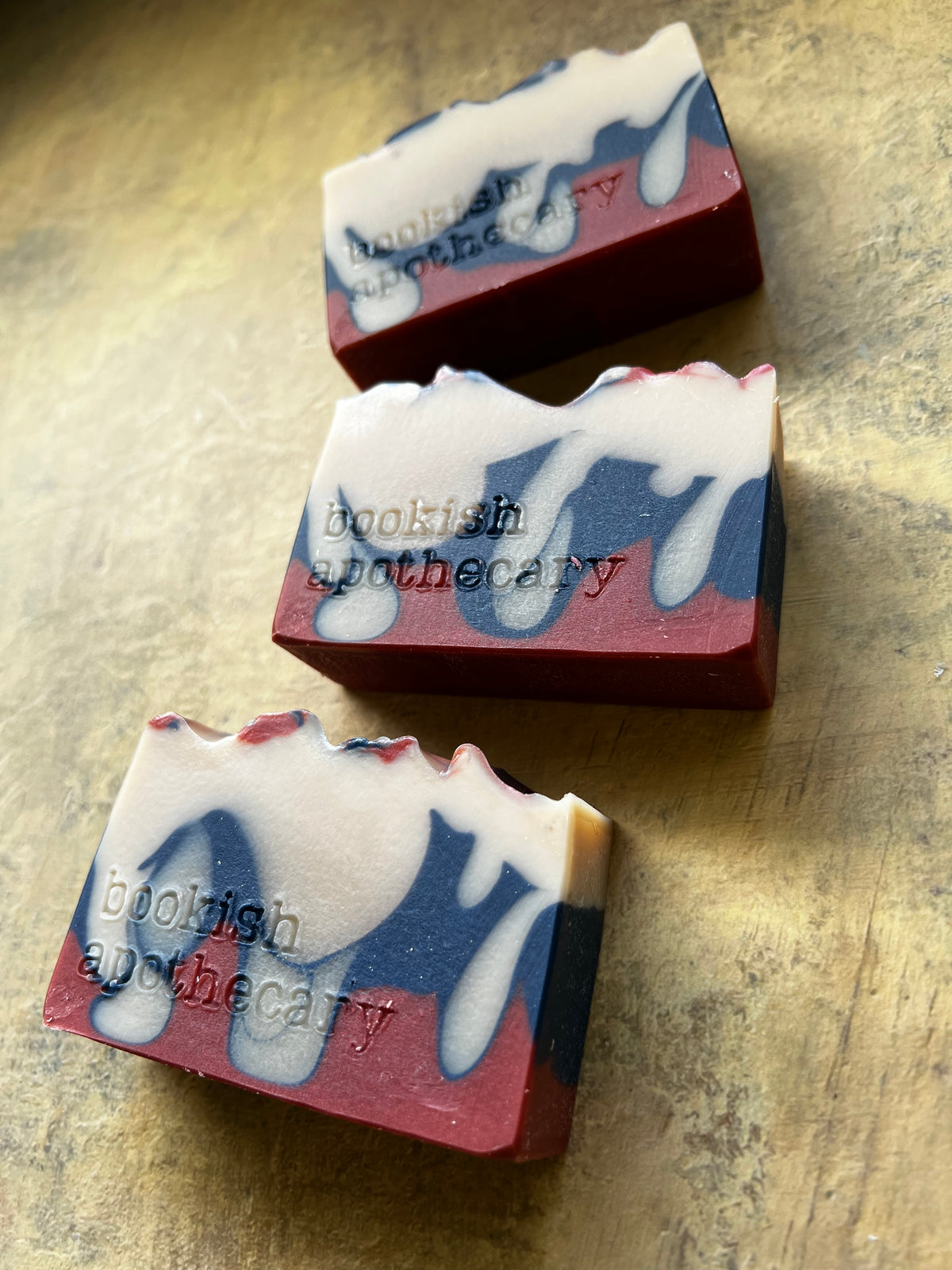 Three handmade soap bars with a marbled pattern of deep red, navy blue, and white, featuring the brand name 'bookish apothecary' embossed on the side, arranged diagonally on a textured gold surface.