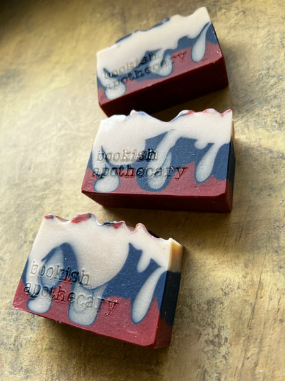Three handmade soap bars with a marbled pattern of deep red, navy blue, and white, featuring the brand name 'bookish apothecary' embossed on the side, arranged diagonally on a textured gold surface.