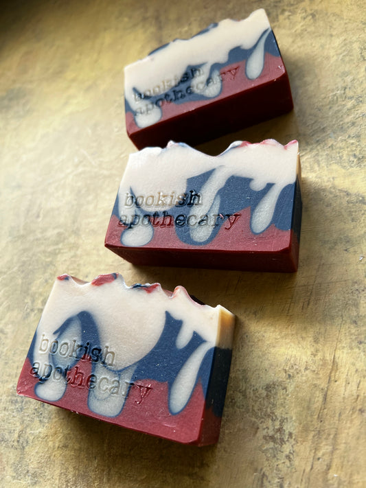 Three handmade soap bars with a marbled pattern of deep red, navy blue, and white, featuring the brand name 'bookish apothecary' embossed on the side, arranged diagonally on a textured gold surface.