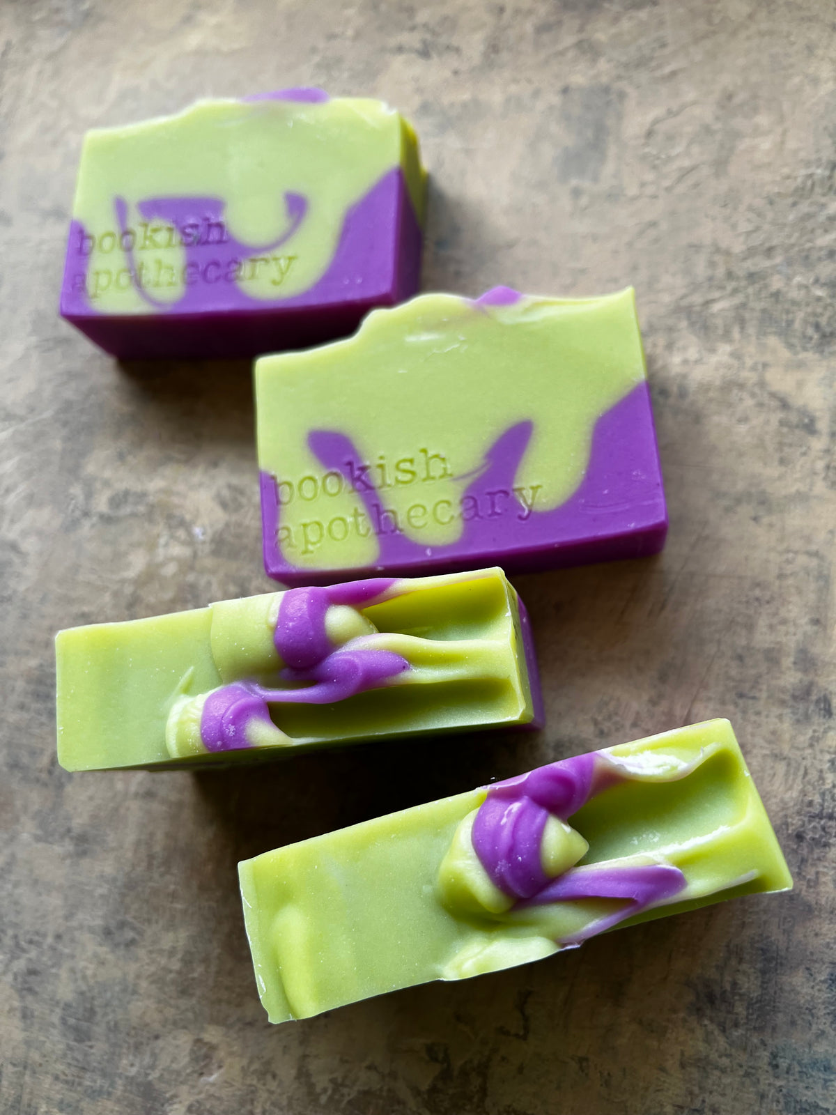 Four rectangular bars of handmade soap with a light green and purple marbled swirl pattern, arranged diagonally on a textured brown surface. The brand name 'bookish apothecary' is embossed on the side of the soaps.