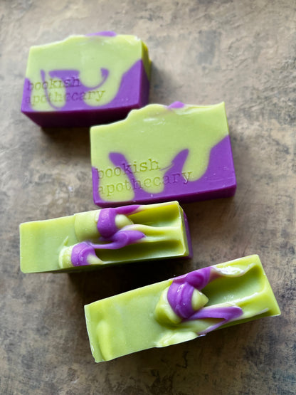 Four rectangular bars of handmade soap with a light green and purple marbled swirl pattern, arranged diagonally on a textured brown surface. The brand name 'bookish apothecary' is embossed on the side of the soaps.