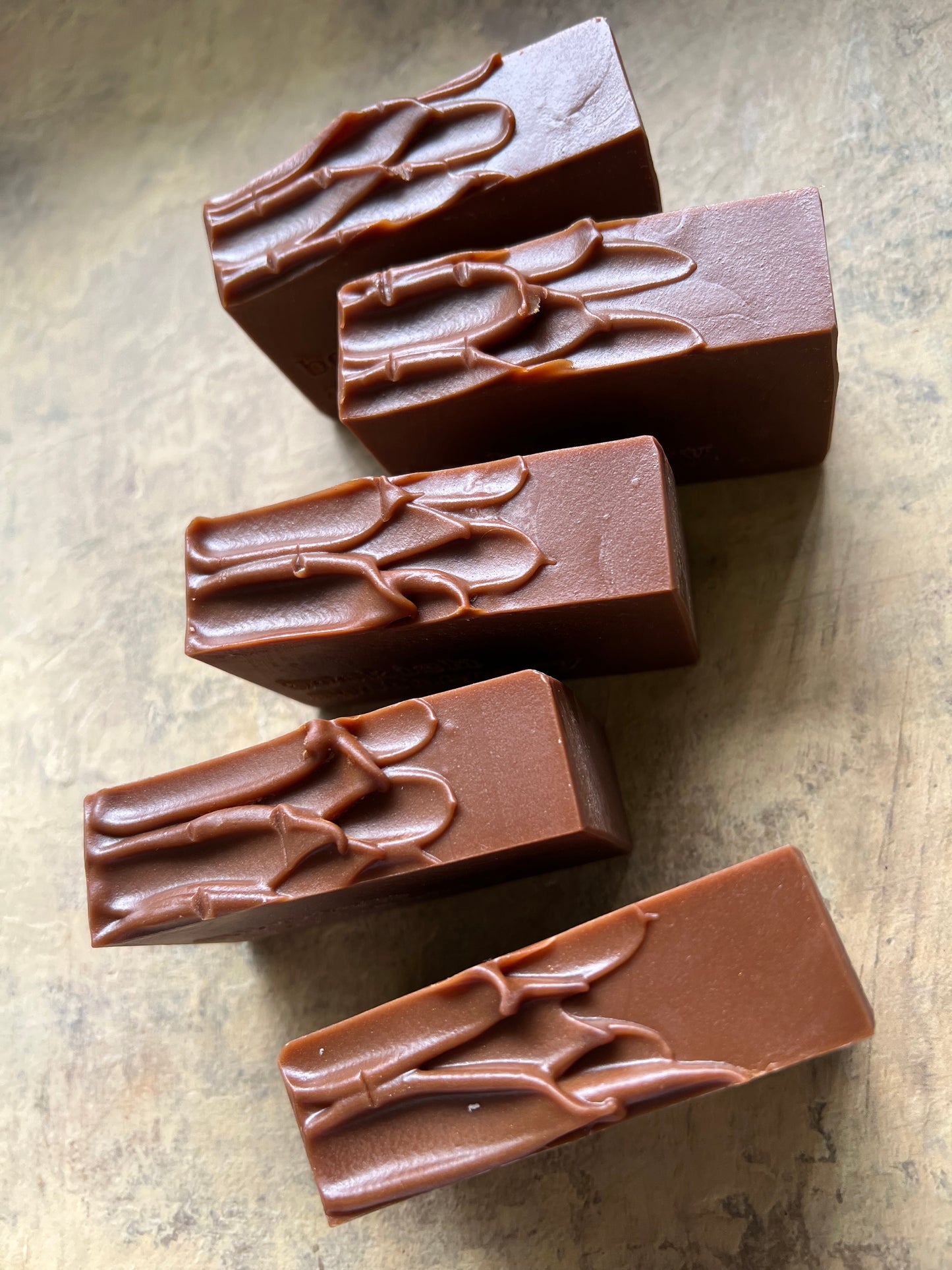 Five rectangular pieces of dark brown chocolate with embossed leaf patterns, arranged on a textured beige surface.