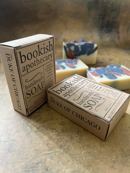 Three bars of handmade soap with vintage-style illustrated packaging, arranged on a textured stone surface. The packaging features a beige background with black typography reading 'Bookish Apothecary'