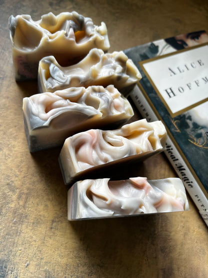 Five decorative handmade soap bars with marbled cream, gray, and pale pink swirls are arranged diagonally on a textured brown surface next to a book.
