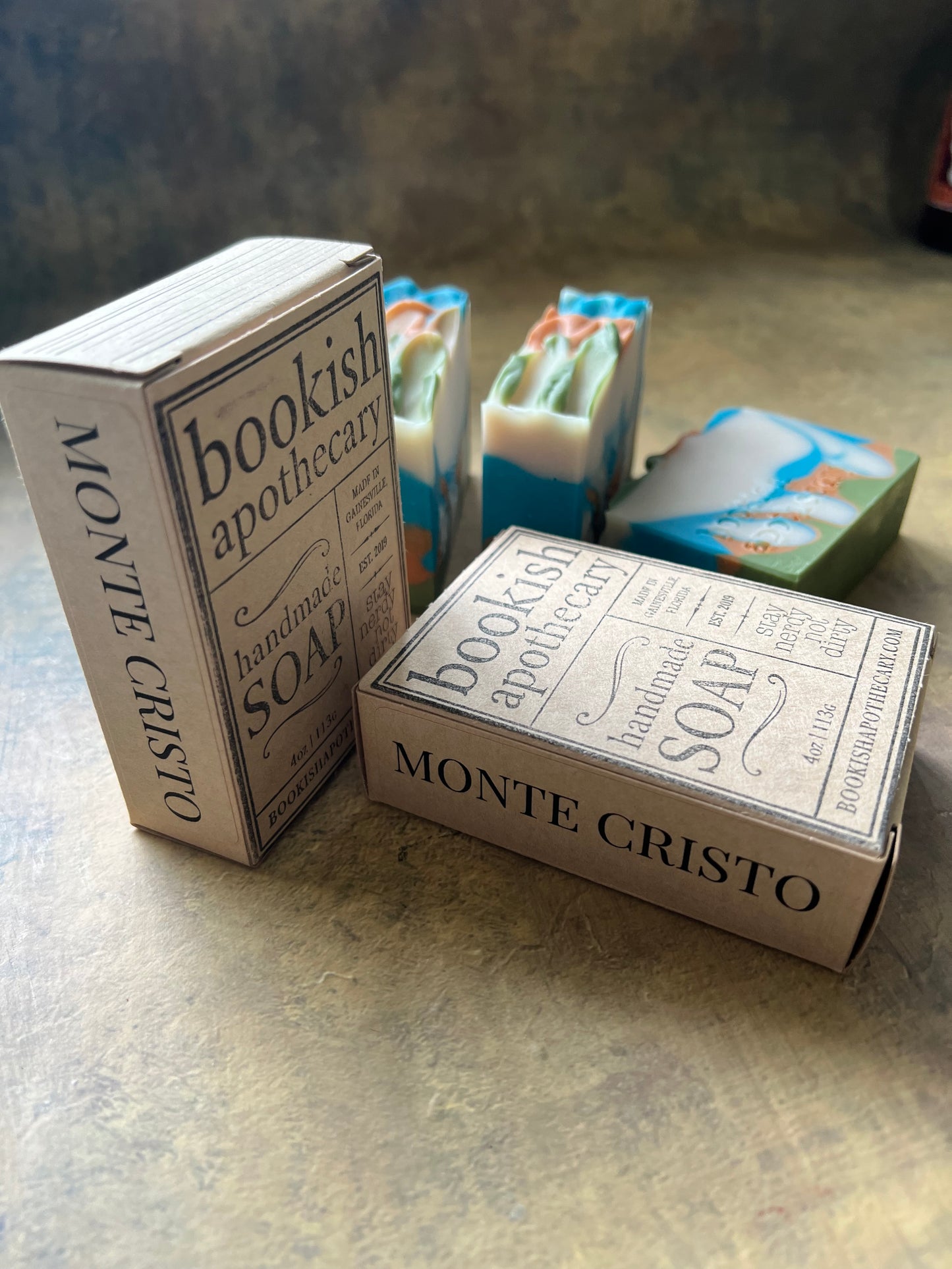 Two bars of handmade bookish apothecary soap in decorative paper packaging, stacked on a textured gray surface.