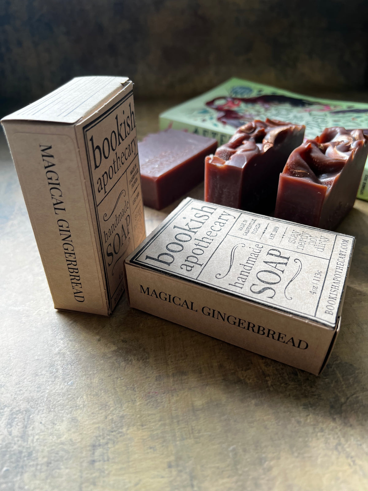 Handmade bar soap in a rustic kraft paper box labeled 'bookish apothecary' and 'MAGICAL GINGERBREAD', shown alongside two dark reddish-brown soap bars on a textured surface.