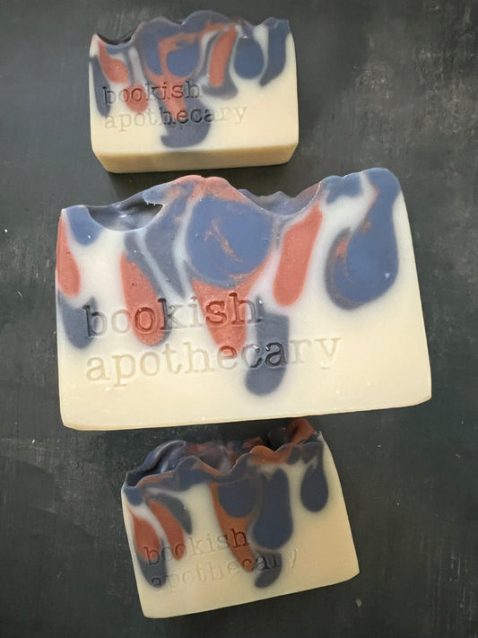 Three bars of swirled soap bars in terracotta and blueish gray stamped with 'bookish apothecary' arranged on a textured gray surface.