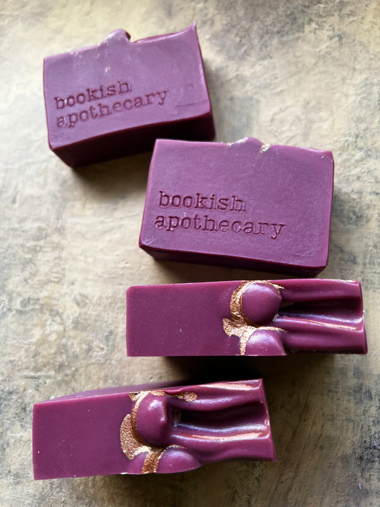 Four purple rectangular bars of soap with the brand name 'bookish apothecary' embossed on the top, arranged on a textured gray surface. Two bars feature a decorative gold foil accent on the side.