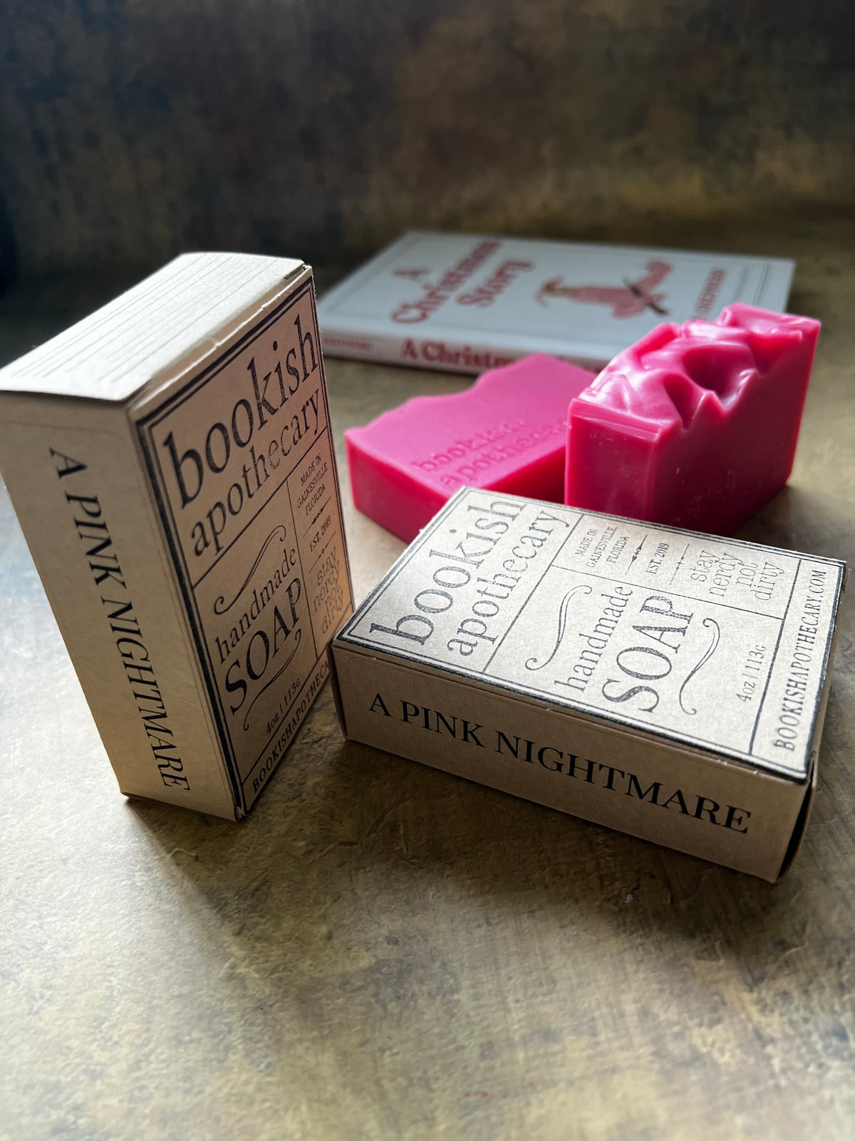 Handmade soap bars and matching paper boxes from Bookish Apothecary, featuring a pink soap bar and vintage-style typography on a rustic bronze surface.