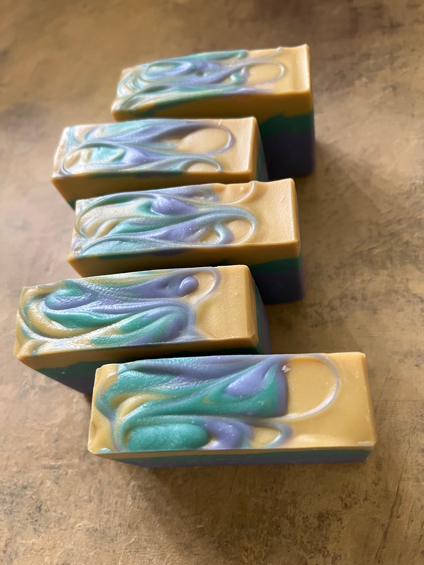 Five rectangular handmade soap bars with swirling teal, lavender, and cream patterns, arranged diagonally on a textured brown surface.