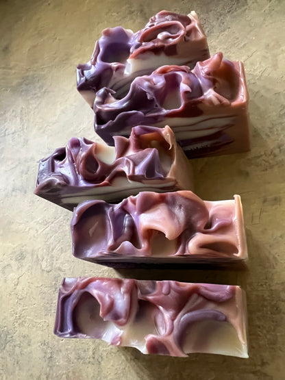 Five rectangular bars of artisanal soap with organic, marbled swirls of purple, mauve, and cream, arranged on a textured beige surface.