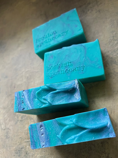 Four handmade rectangular bars of soap in shades of turquoise, teal, and blue with purple marbling, arranged on a textured beige surface. Two bars feature stamped text reading 'rootish apothecary'.