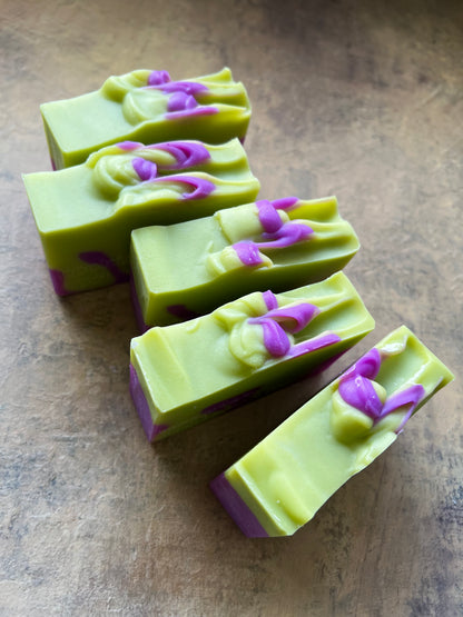 Five rectangular bars of handmade soap arranged diagonally on a textured brown surface. The soap is primarily a light lime green with purple swirls and piped decorative detailing on top.