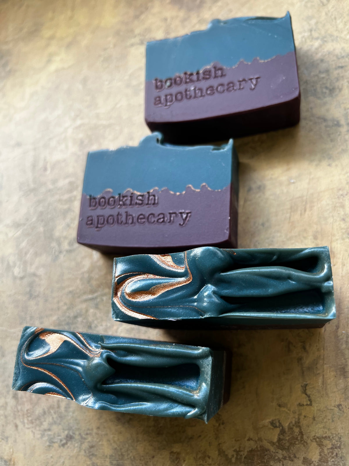 Four artisanal soap bars from Bookish Apothecary arranged diagonally on a textured beige stone surface. The soaps feature a rustic dual-tone design with a deep teal blue lower half and a muted purple upper half, with one bar showcasing a copper swirl pattern.