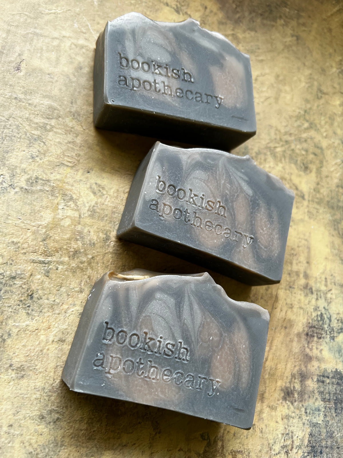 Three handmade soap bars with wavy edges, featuring a dark gray and brown marbled pattern and embossed text reading 'bookish apothecary', arranged vertically on a textured, rustic gold-toned surface.