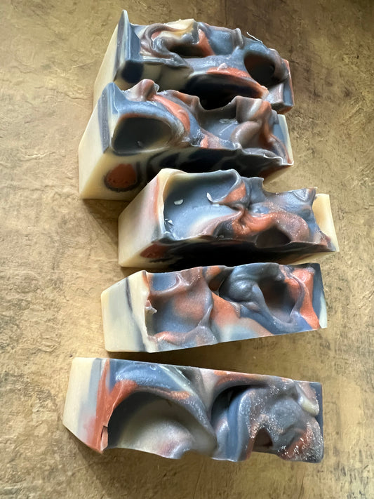 Five handmade marbled soap bars with a swirled pattern of slate blue, terracotta orange, cream, and dark gray, arranged vertically on a textured tan surface.