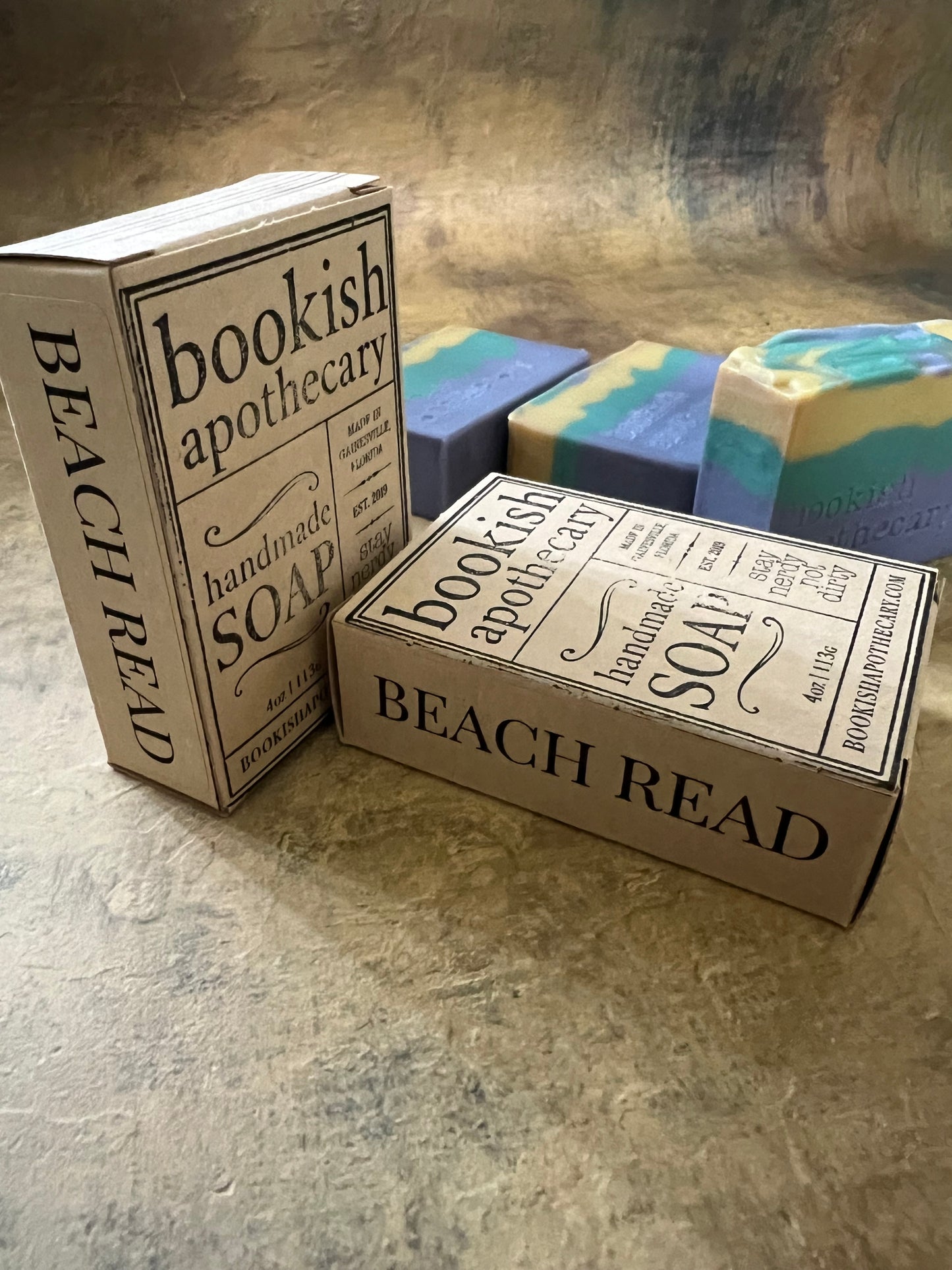 Three rectangular bars of handmade soap in pastel blue, green, and cream, each in its own decorative cardboard box. The boxes are styled like vintage book covers with black typography reading 'Bookish Apothecary' and 'BEACH READ'.