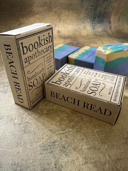 Three rectangular bars of handmade soap in pastel blue, green, and cream, each in its own decorative cardboard box. The boxes are styled like vintage book covers with black typography reading 'Bookish Apothecary' and 'BEACH READ'.
