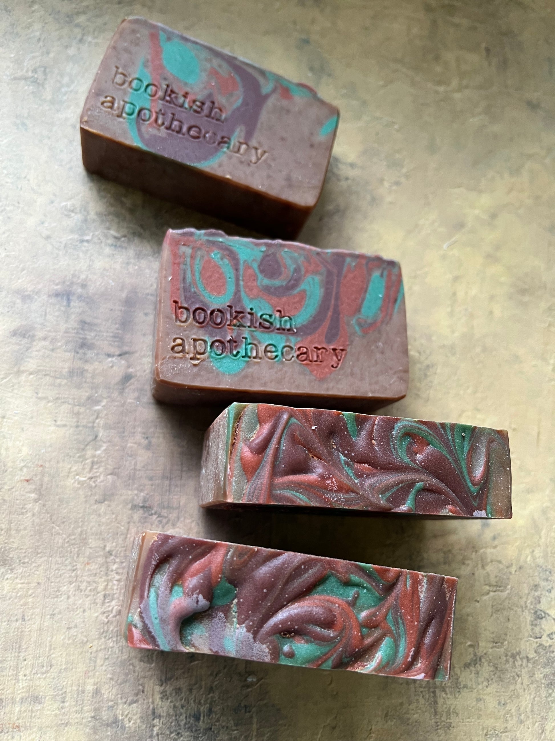 Four handmade artisanal soap bars with a marbled pattern of brown, teal, and red, each stamped with the brand name 'bookish apothecary', arranged on a textured gray surface.