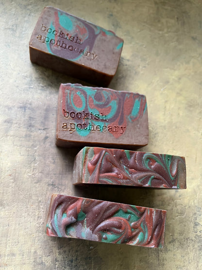 Four handmade artisanal soap bars with a marbled pattern of brown, teal, and red, each stamped with the brand name 'bookish apothecary', arranged on a textured gray surface.
