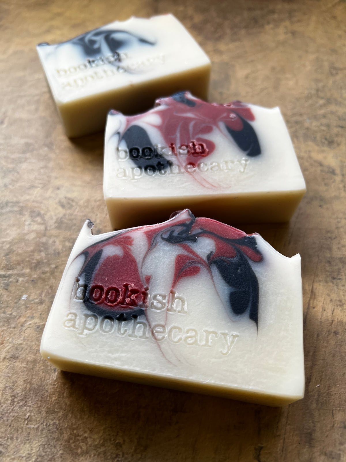 Three handmade artisanal soap bars with a cream base and red, black, and white marbled swirls, embossed with the brand name 'bookish apothecary', arranged diagonally on a rustic wooden surface.