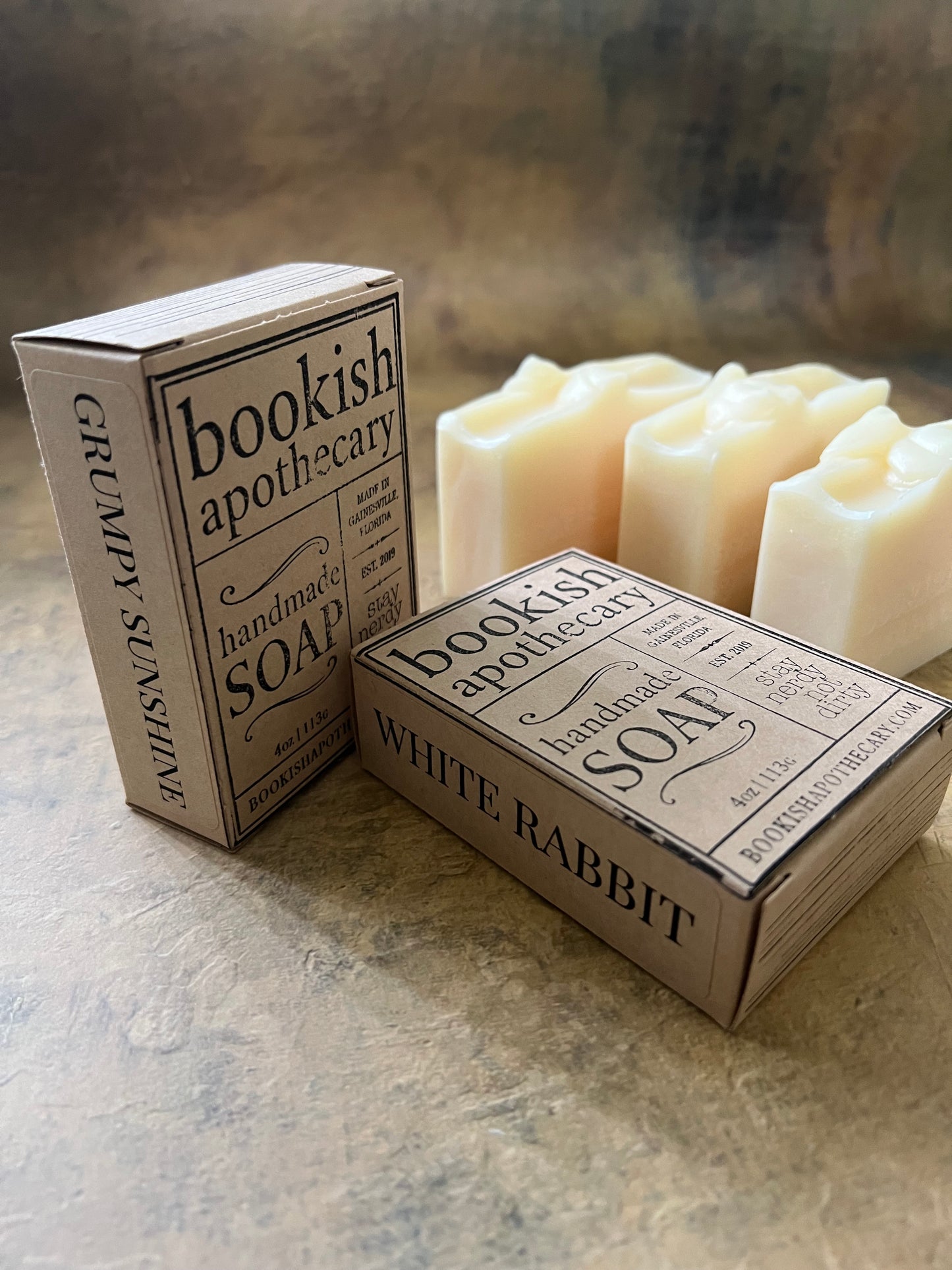 Two boxed bars of handmade soap on a textured stone surface, with three cream-colored soap bar partially visible behind the boxes. The boxes are labeled 'Bookish Apothecary' and show scents 'White Rabbit'